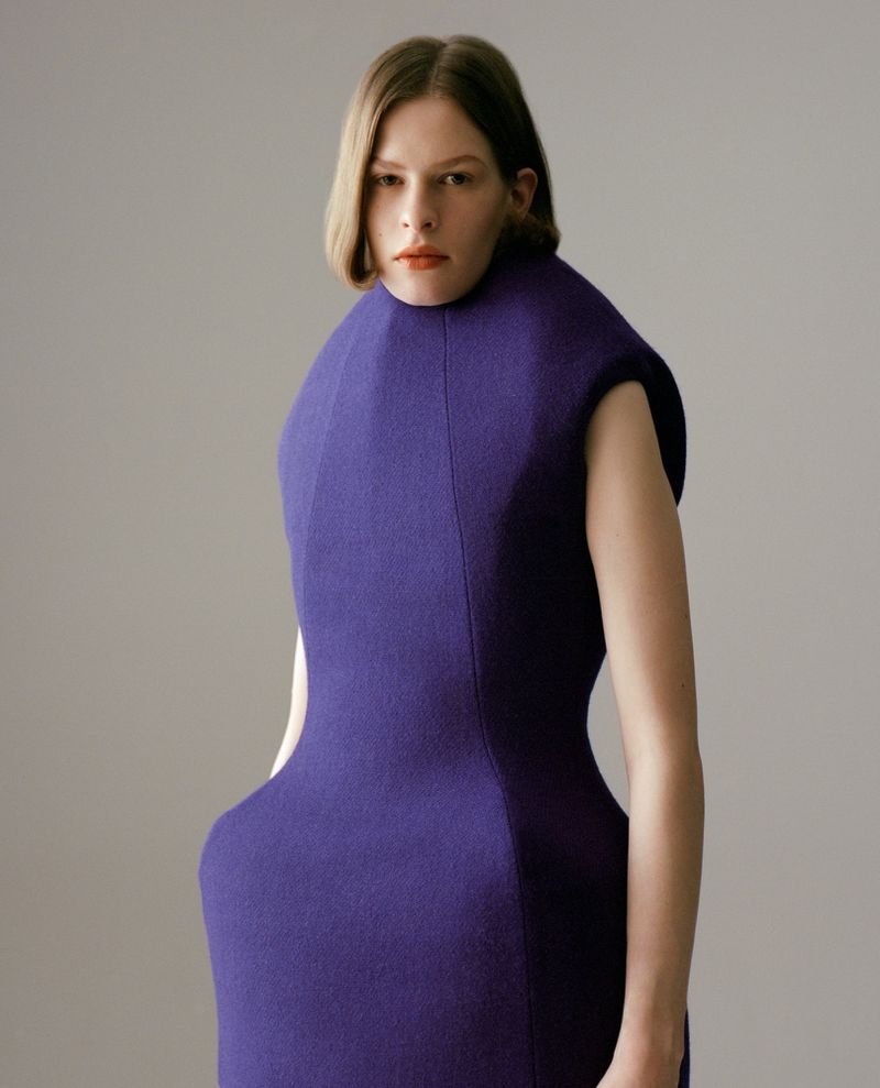 Model Hannah Weber wearing a sculptural structured purple dress against a neutral grey studio backdrop.