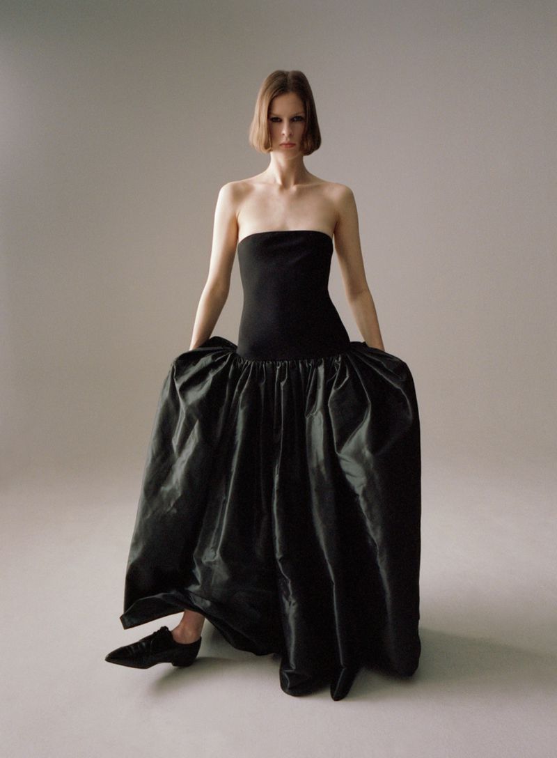 Model Hannah Weber posing in a strapless black gown with an exaggerated voluminous skirt in a minimalist studio setting.