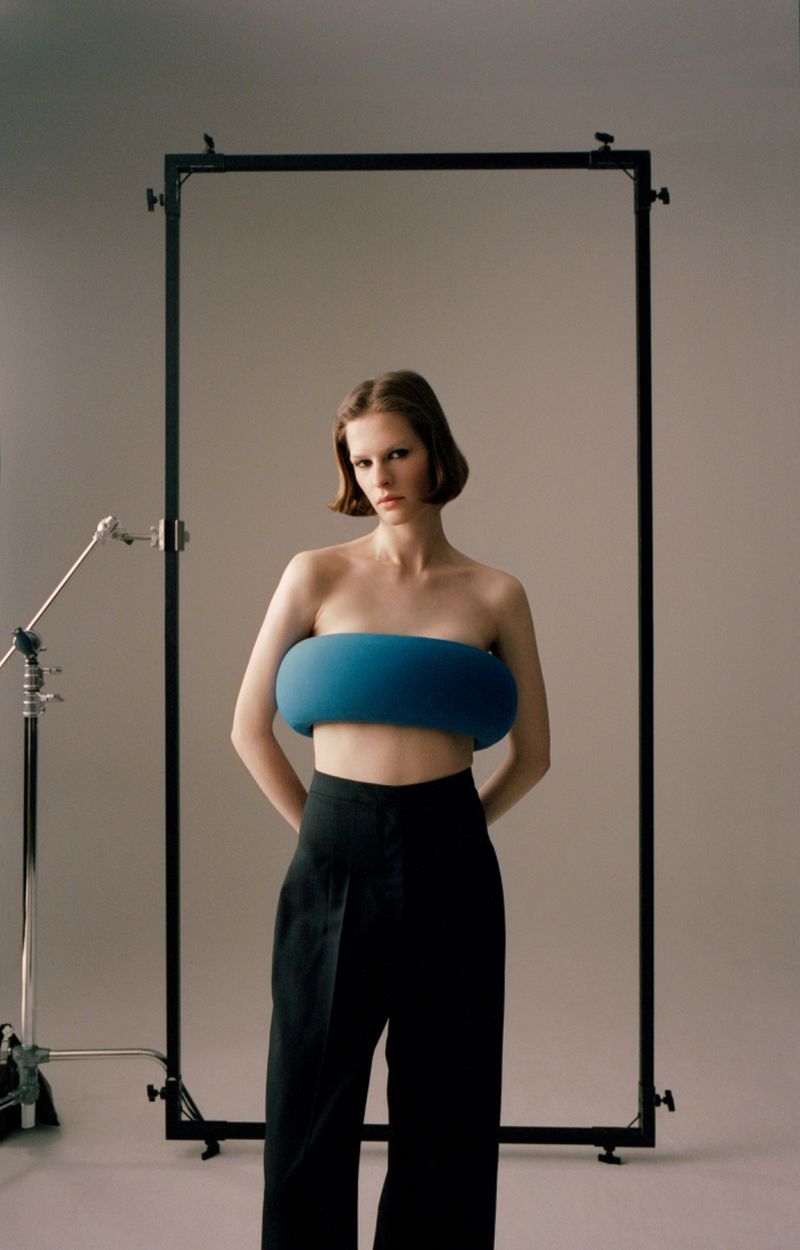 Model Hannah Weber wearing a sculptural blue bandeau top and black trousers framed by studio equipment against a grey backdrop.