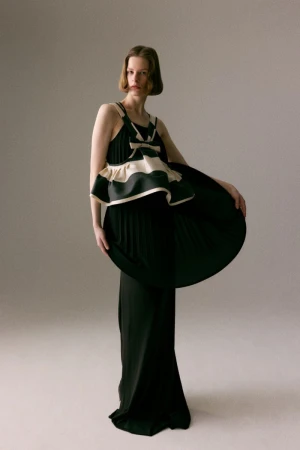 Model Hannah Weber poses in a floor-length black pleated gown with a sculptural cream-and-black top against a neutral studio backdrop.