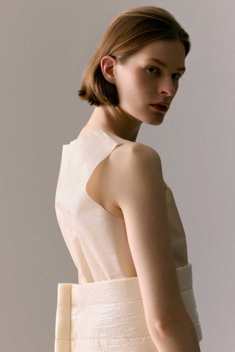 Model Hannah Weber looks over her shoulder wearing a structured cream sleeveless top against a soft neutral studio backdrop.