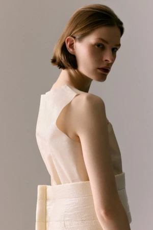Model Hannah Weber looks over her shoulder wearing a structured cream sleeveless top against a soft neutral studio backdrop.