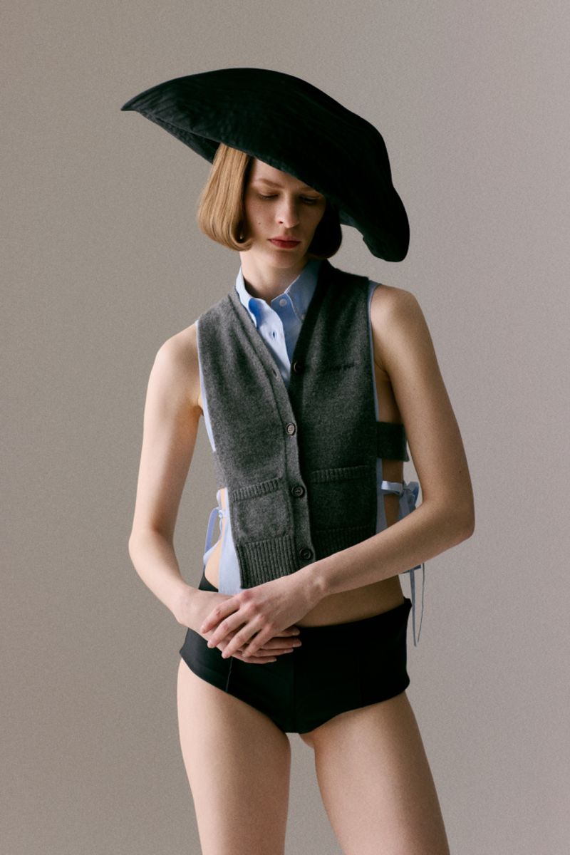 Model Hannah Weber in an oversized black sculptural hat and grey knit vest against a neutral studio backdrop.