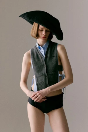 Model Hannah Weber in an oversized black sculptural hat and grey knit vest against a neutral studio backdrop.