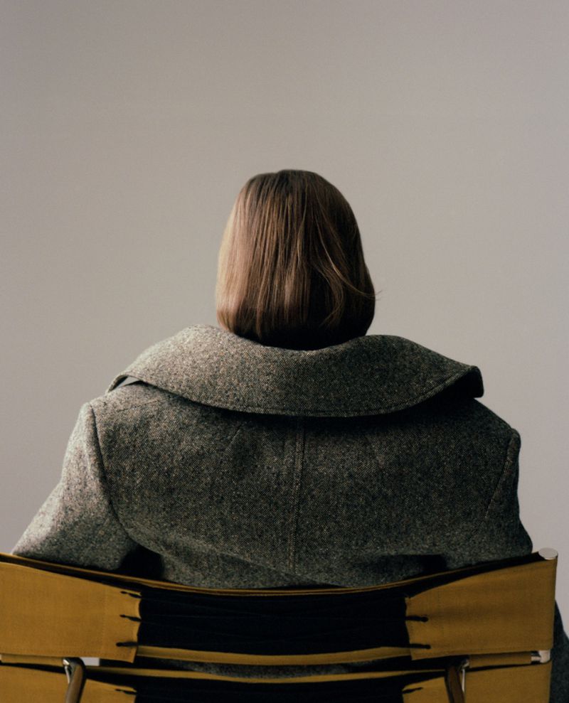 Model Hannah Weber viewed from behind wearing a heavy grey tweed coat while seated in a chair against a neutral studio backdrop.