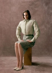 Model Hannah Weber seated on a wooden block wearing a padded beige button-up shirt and green lace pencil skirt against a textured backdrop.