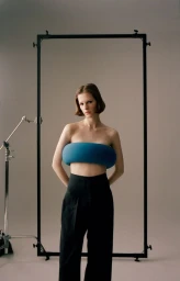 Model Hannah Weber wearing a sculptural blue bandeau top and black trousers framed by studio equipment against a grey backdrop.