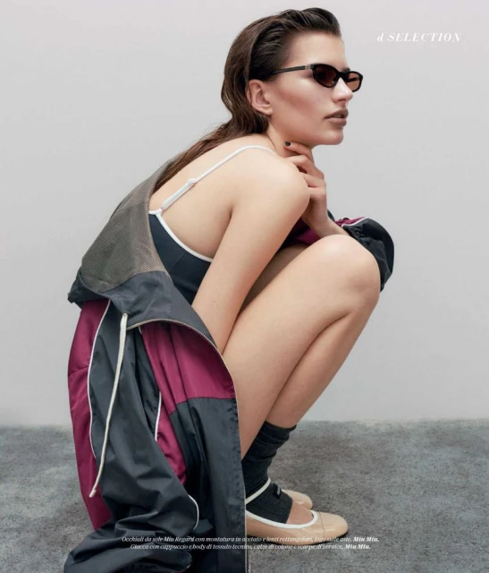 Woman in sunglasses and swimsuit crouches indoors, wearing jacket with grey, black and maroon panels, and ballet shoes with socks.