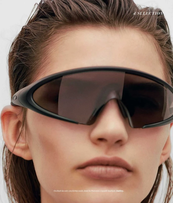 Close-up portrait of a model wearing dark futuristic shield sunglasses and wet-look hair against a neutral background.