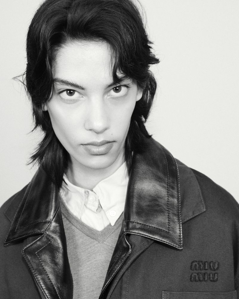 Monochromatic close-up portrait of a model with dark wavy hair wearing a jacket with leather lapels and an embroidered chest logo.
