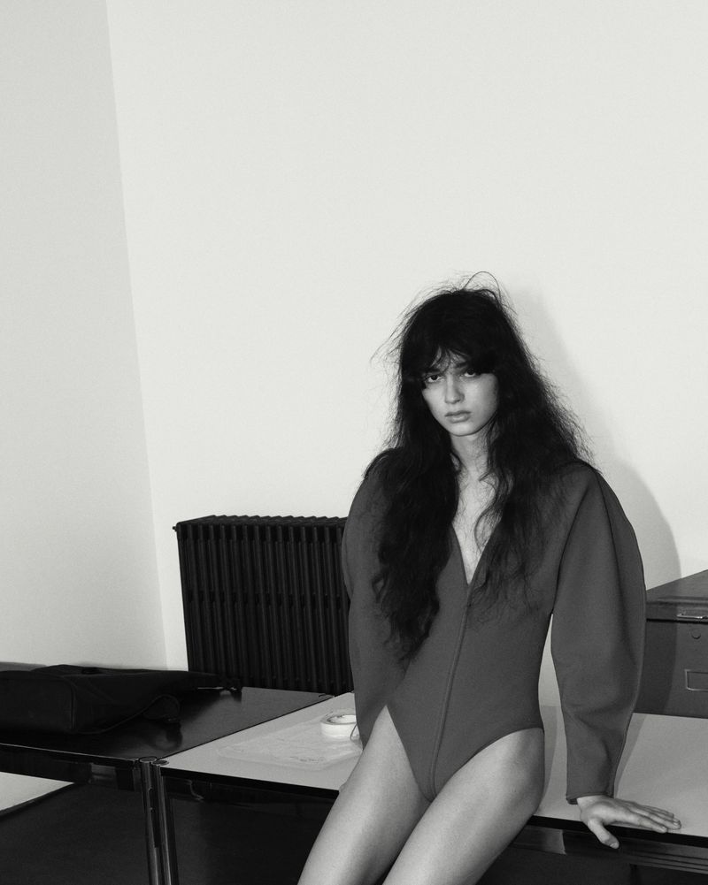 Model with long dark hair wearing a sculptural long-sleeve bodysuit while seated on a desk against a plain wall.