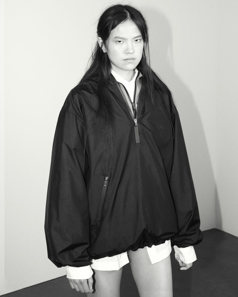 Monochrome portrait of an East Asian model wearing an oversized black zip-up pullover over a white shirt against a neutral wall.
