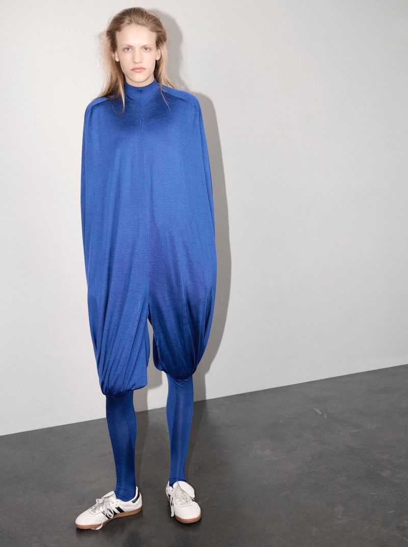 A model standing in an oversized draped royal blue jumpsuit with matching blue tights and white sneakers against a neutral wall.