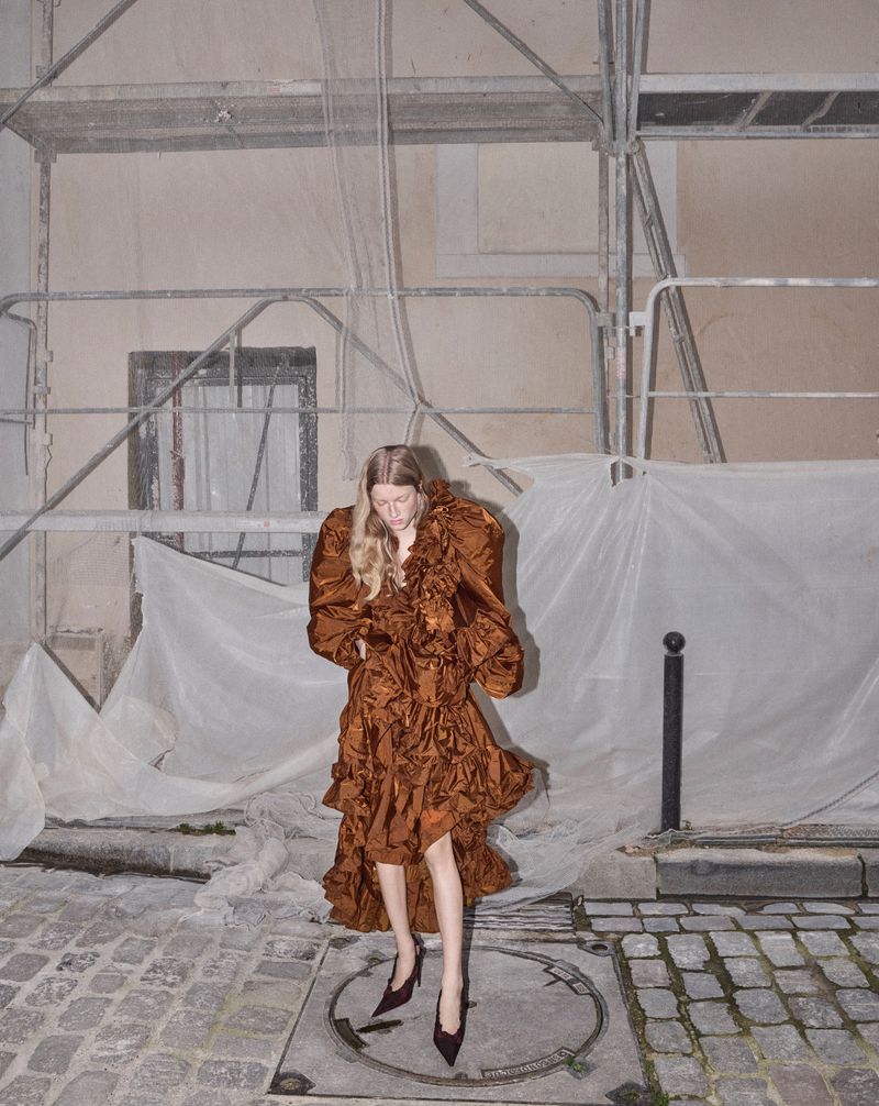 Model wearing a voluminous brown ruffled dress and pointed high heels standing before scaffolding on a cobblestone street.