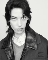 Monochromatic close-up portrait of a model with dark wavy hair wearing a jacket with leather lapels and an embroidered chest logo.
