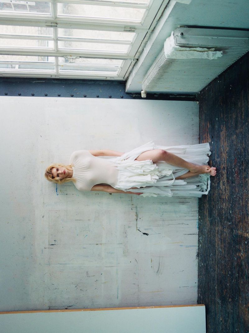 Camille Chifflot in a layered off-white ruffle dress leaning against a distressed wall next to windows.