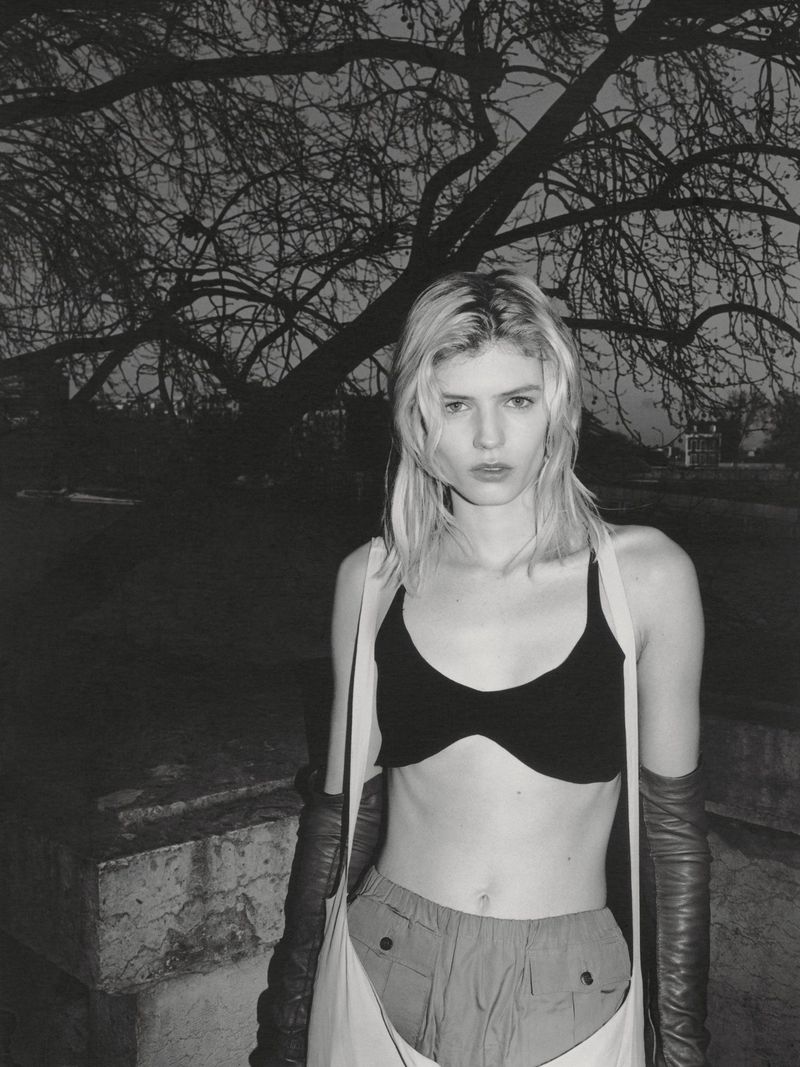 Monochrome portrait of a female model wearing a crop top and gloves outdoors beneath dark tree branches.