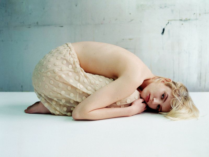 Model curled up on a white surface wearing a sheer dotted skirt with a pale wall background