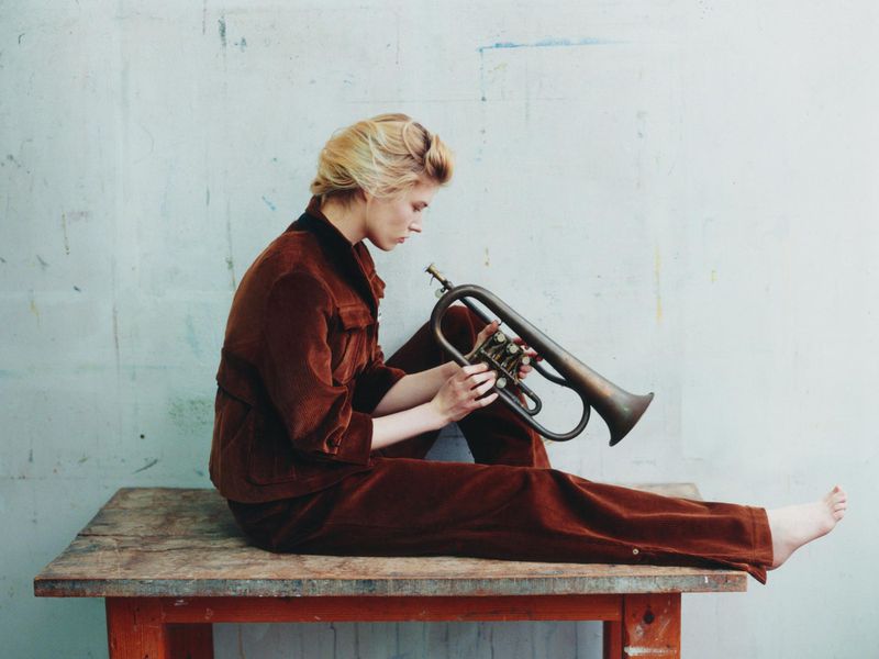 Model in a brown corduroy suit seated barefoot on a wooden table holding a brass flugelhorn against a textured backdrop.