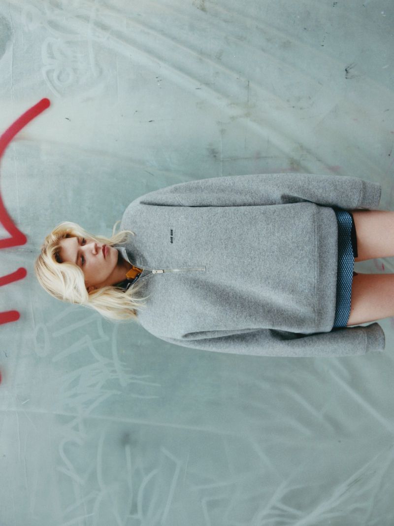 Model Camille Chifflot in a grey half-zip fleece pullover standing against a translucent wall with red graffiti markings.