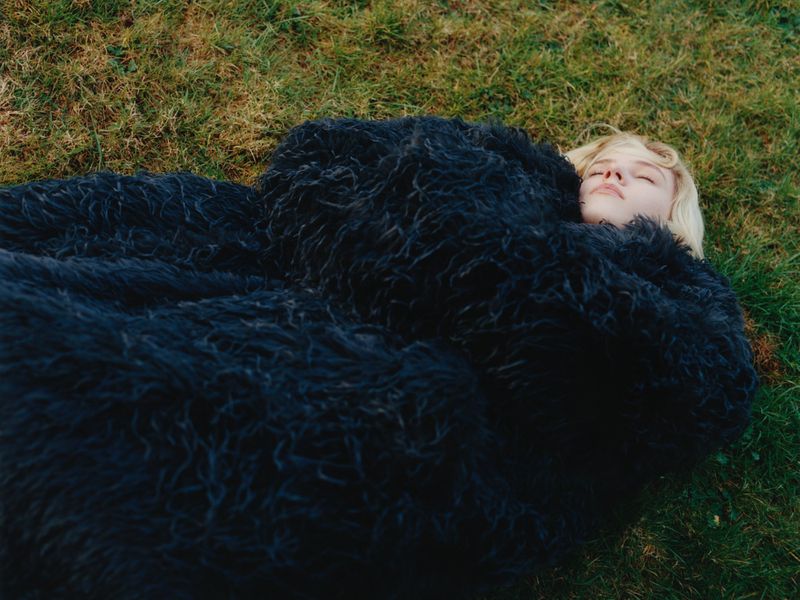 Model Camille Chifflot lying on green grass bundled in an oversized shaggy black coat with her eyes closed.