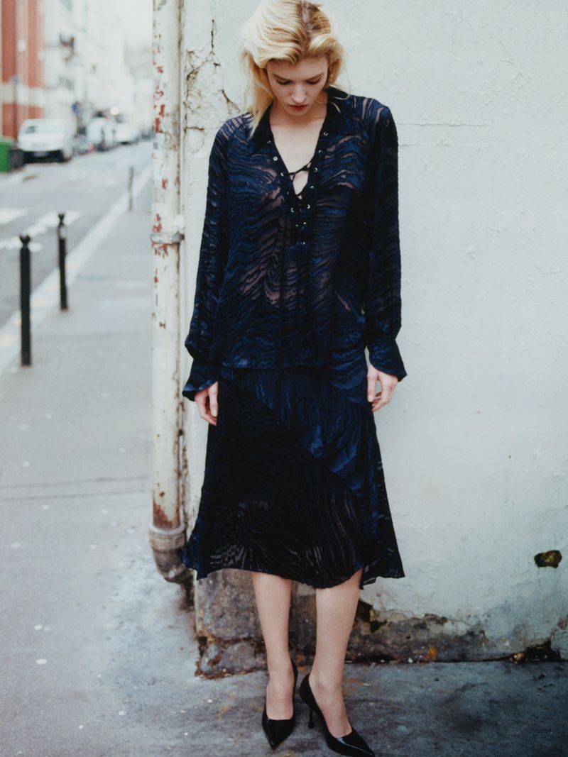 Model Camille Chifflot wearing a sheer patterned dark navy outfit and black heels while standing against a wall on a city sidewalk.