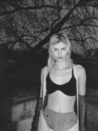 Monochrome portrait of a female model wearing a crop top and gloves outdoors beneath dark tree branches.