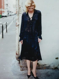 Model Camille Chifflot wearing a sheer patterned dark navy outfit and black heels while standing against a wall on a city sidewalk.