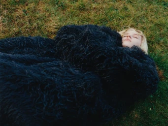 Model Camille Chifflot lying on green grass bundled in an oversized shaggy black coat with her eyes closed.