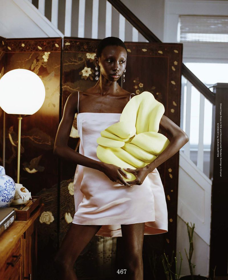 Model Alay Deng poses indoors in a pale blush satin mini dress, cradling a large yellow sculptural object beside an ornate screen.