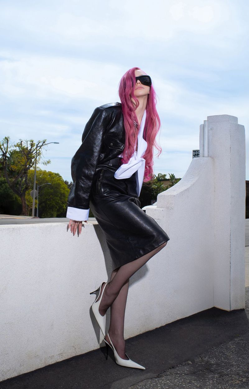 Adéla Jergová with pink hair wearing a black leather skirt suit, sunglasses, and white heels leaning on a white wall.