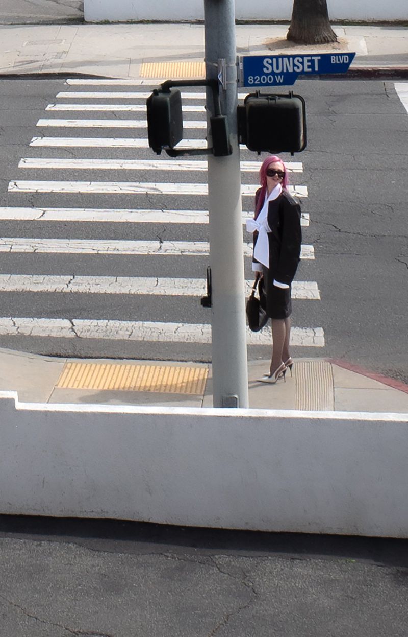 Adéla Jergová standing next to a Sunset Blvd pedestrian crosswalk pole in a black blazer, white bow top, and heels.
