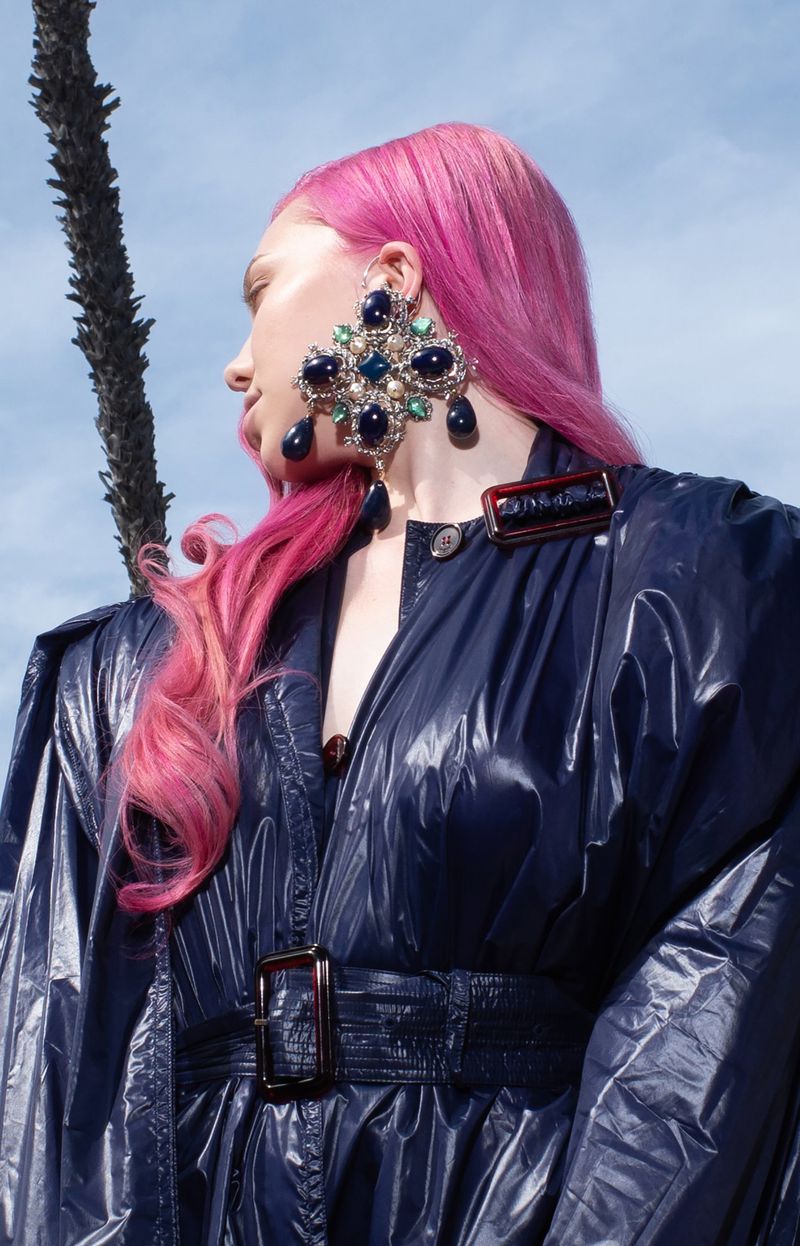 Adéla Jergová with bright pink hair wearing a large ornate jeweled earring and a glossy belted navy coat against an outdoor sky.