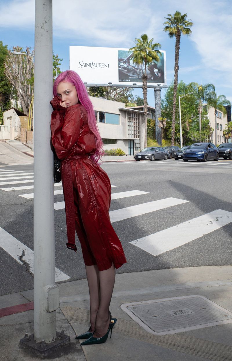 Adéla Jergová with bright pink hair wearing a shiny red belted trench coat and high heels while leaning against a street lamppost.