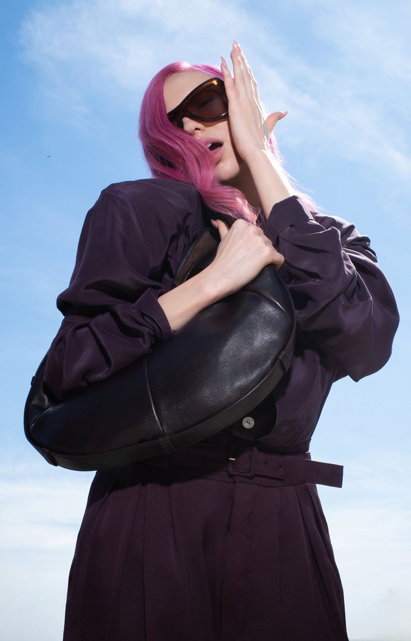 Adéla Jergová with bright pink hair wearing dark sunglasses and a plum outfit holding a black handbag under a clear blue sky