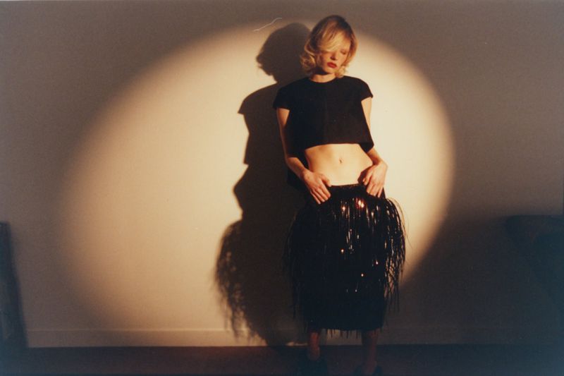 Model standing under a dramatic spotlight in front of a plain wall, wearing a black cropped top and dark fringed skirt.