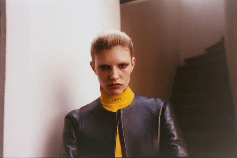 Model Camille Chifflot wearing a yellow Loewe turtleneck and black leather jacket in a dimly lit hallway.
