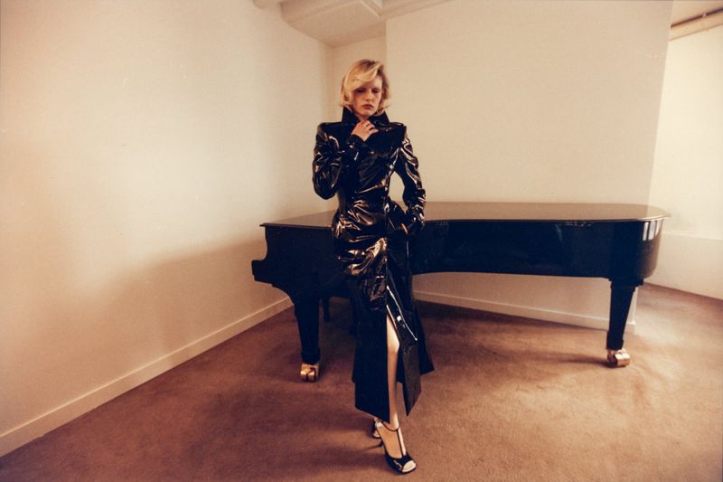 Model Camille Chifflot wearing a glossy black trench coat posed beside a grand piano in a minimalist room.