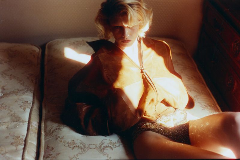 Camille Chifflot reclining on a patterned mattress wearing a caramel leather jacket and embellished briefs in direct sunlight.