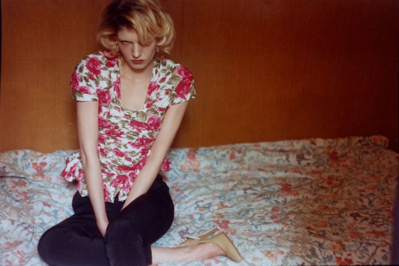 Model Camille Chifflot in a floral short-sleeved top and black trousers seated on a floral bedspread.