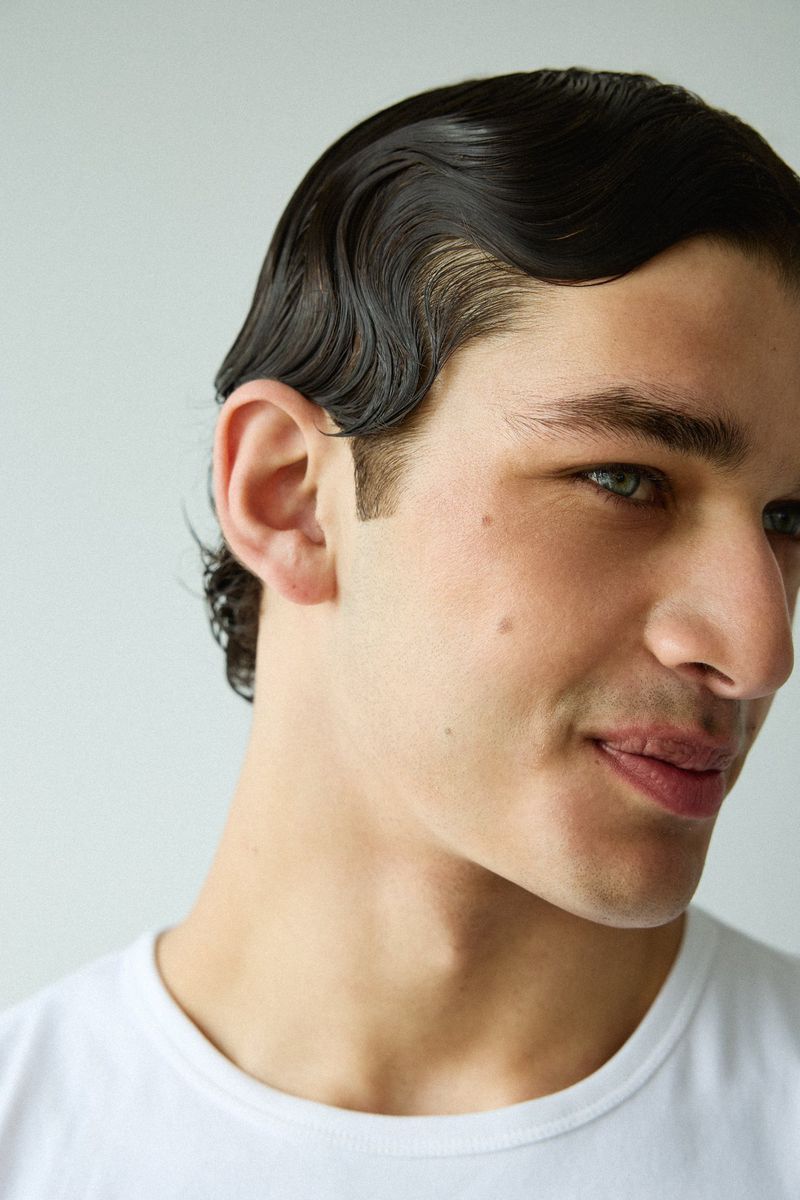 Close-up profile portrait of male model Pablo Fernandez with slicked finger wave hair wearing a white t-shirt against a plain backdrop.
