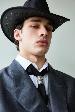 Male model wearing a black cowboy hat, grey blazer, white shirt, and black scarf with a silver ring against a plain studio background.