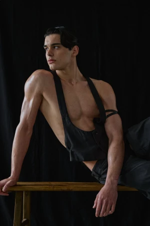 Shirtless male model Pablo Fernandez posing in a black harness vest and trousers on a wooden bench against a black backdrop.