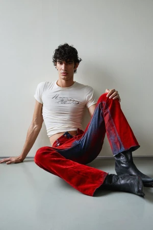 Model Pablo Fernandez seated on a studio floor wearing a white graphic t-shirt, two-tone red and blue flare trousers, and black boots.