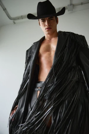 Model wearing a black cowboy hat and an open black leather fringe coat against a white studio wall.