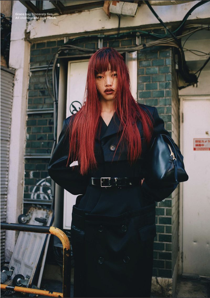 Model Yuki Beniya with vibrant red hair wearing a belted black trench coat against a tiled exterior wall.
