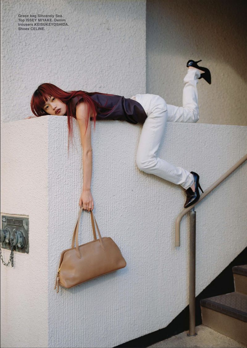 Model Yuki Beniya lying across a textured white wall ledge while holding a tan leather handbag for DEW Magazine.
