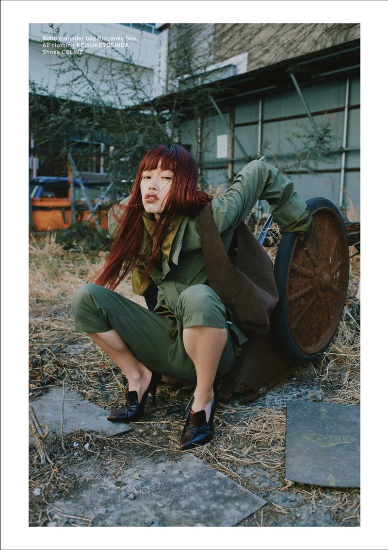 Model Yuki Beniya with red hair crouching in an olive green utility jumpsuit and black heels in an industrial yard.
