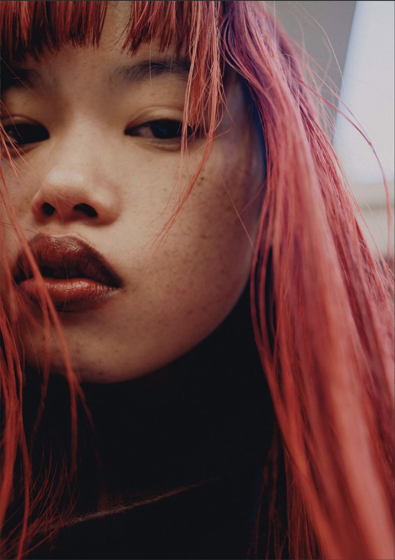 Close-up portrait of an East Asian model with bright red hair, bangs, and freckles looking directly at the camera.