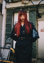 Model Yuki Beniya with vibrant red hair wearing a belted black trench coat against a tiled exterior wall.